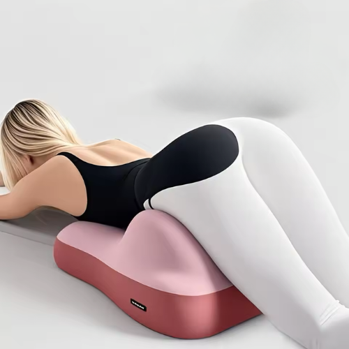 A woman in a black and white bodysuit lies face down on the Todle Couple Pillow, a pink wedge designed to support her hips and legs, against a minimal, light background.