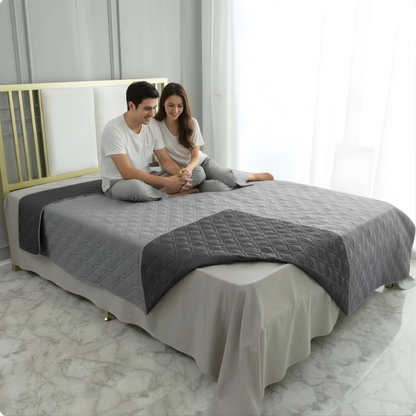 A smiling couple in matching outfits sits cross-legged on a neatly made bed with gray bedding, featuring the My Store Reversible Waterproof Bed Blanket – Discreet Comfort for Couples, in a bright room with large windows and marble floors.